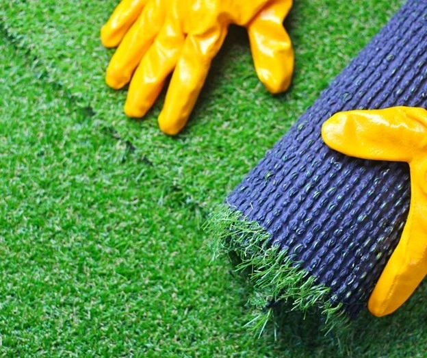 The image features lawn maintenance by Hawkins Landscaping, showcasing the installation of artificial turf with yellow gloves handling the process, emphasizing a low-maintenance and sustainable lawn solution.