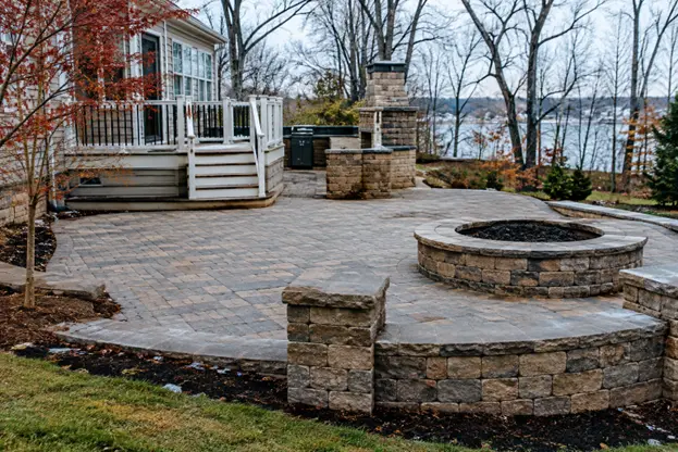 A spacious paver patio with a circular stone fire pit, curved retaining wall, and outdoor stone fireplace showcases masonry hardscape features by Hawkins Landscaping for a durable outdoor gathering space.