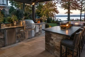 A covered outdoor kitchen with a stainless-steel grill, stone countertops, built-in refrigerator, and bar seating overlooks a waterfront patio, showcasing outdoor kitchen services by Hawkins Landscaping for comfortable fall entertaining.
